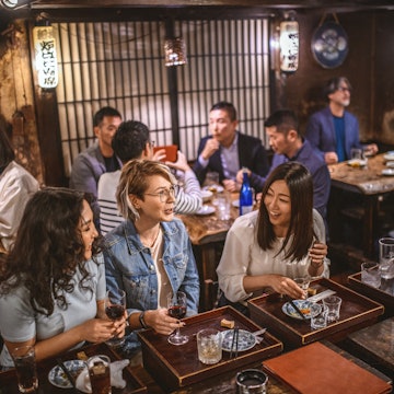 Wide angle view of Mongolian, Japanese, and Caucasian women sitting at sushi bar and other patrons in Tokyo izakaya.